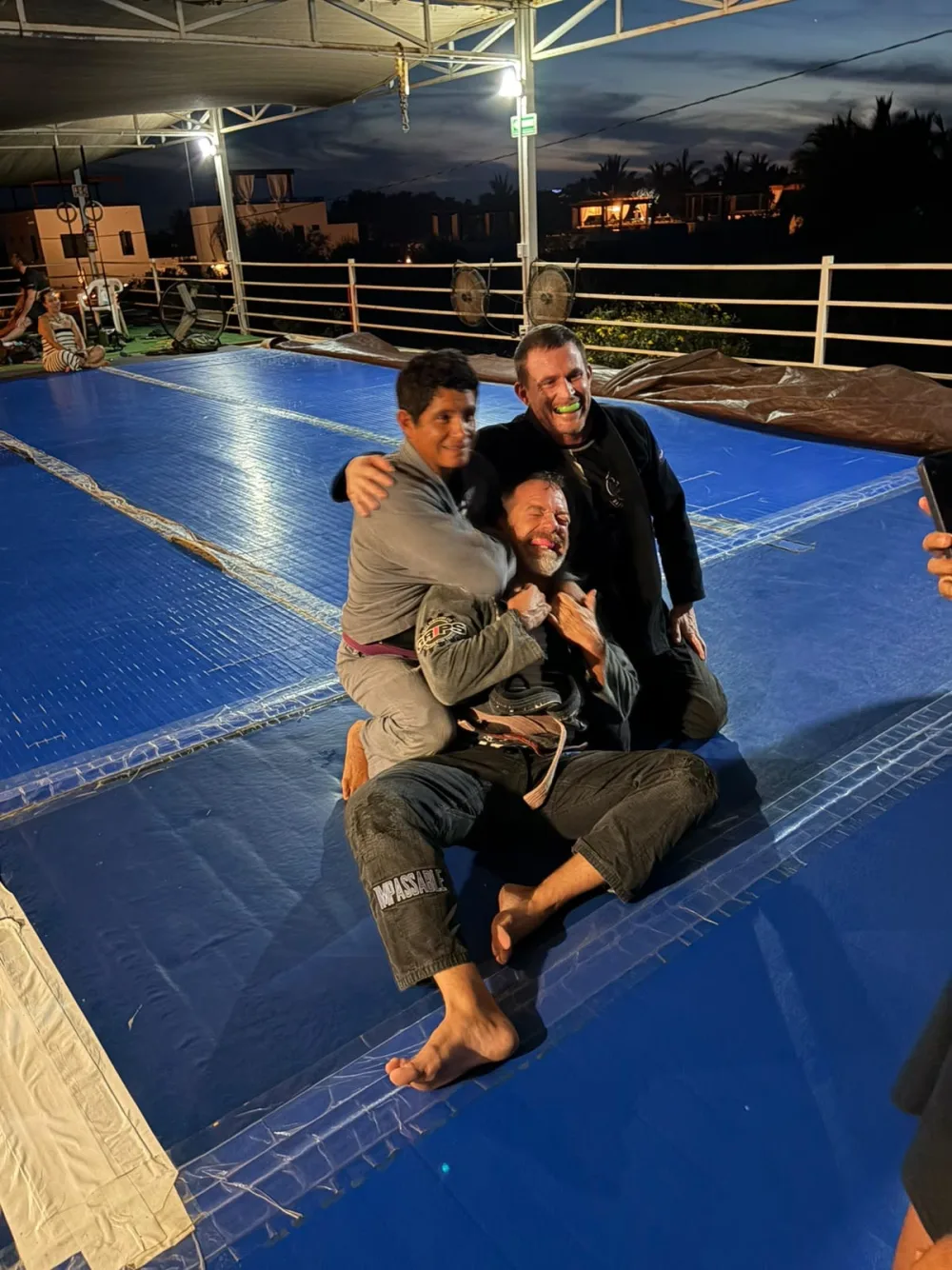 Three Todos Santos BJJ members laughing together on the blue training mats
